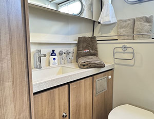 Avalon Bad Heckkabinekabine, Blick auf Waschtisch, schräg vorne, darüber Spiegel – darin spiegelt sich das seitliche Bullauge. Rechts unten die Toilette