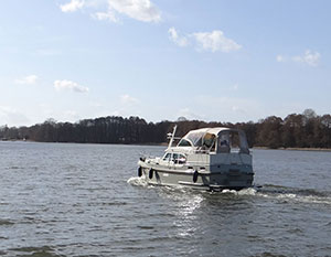 Linssen 30.0 AC Lili in Fahrt - von rechts nach links, Blick von schräg hinten auf die Backbordseite und das Heck, geschlossene Persenning. blauer Himmel mit kleinen Wolken, im Hintergrund bewaldetes Ufer